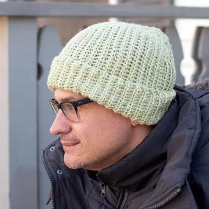 Könnte beinhalten: Eine hellgrüne Strickmütze mit gerippter Struktur. Die Mütze wird von einer Person mit Brille getragen.