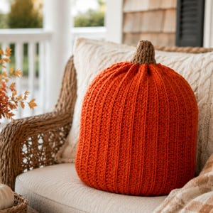 May include: A round, orange, crocheted pumpkin pillow with a textured, ribbed surface and a small, brown, crocheted stem at the top. The pumpkin is crafted from yarn and features a consistent, vertical stitch pattern throughout its rounded body.