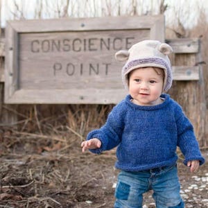 May include: A young child wearing a blue knitted sweater and jeans, with a brown and white furry hat with ear flaps. The child is standing in front of a wooden sign that reads "CONSCIENCE POINT".