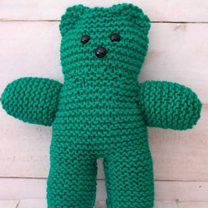 Puede incluir: Un oso de peluche verde tejido a mano, con ojos de botón negros. El oso tiene un diseño clásico con cabeza redonda, orejas pequeñas y brazos extendidos. La lana suave y texturizada le da al oso un aspecto acogedor y artesanal.