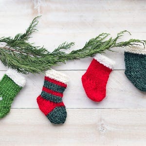 Puede incluir: Cuatro pequeñas medias de Navidad de punto en rojo, verde y blanco. Las medias están colgadas de una rama verde.
