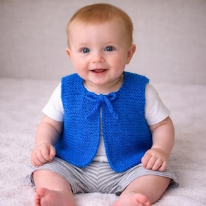 May include: A baby wearing a bright blue knitted vest with a bow closure. The vest is over a white t-shirt and paired with gray and white striped shorts. The baby is smiling, with a soft, neutral background.