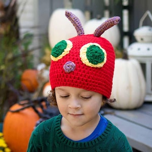 Può includere: Un cappello rosso all'uncinetto con dettagli verdi, gialli e viola, progettato per assomigliare a un bruco. Il cappello ha due antenne viola. Il bambino indossa un maglione verde e una maglietta blu.