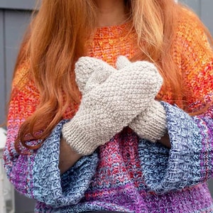 May include: A pair of beige knitted mittens with a textured stitch pattern. The mittens are held together in front of a person wearing a colorful sweater.