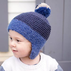 May include: A blue and gray knitted hat with ear flaps and a pom pom on top. The hat is worn by a child.