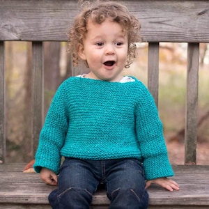 May include: A young child wearing a teal blue knitted sweater with long sleeves and a relaxed fit. The child is sitting on a wooden bench and has curly blonde hair.