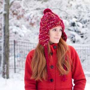 May include: A woman wearing a red knitted hat with ear flaps and a pom pom on top. The hat is a mix of red, pink, and white yarn. She is also wearing a red coat.