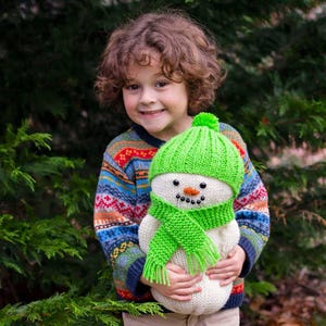 May include: A child with curly brown hair is holding a knitted snowman toy. The snowman is wearing a green hat and scarf. The child is wearing a colorful sweater.