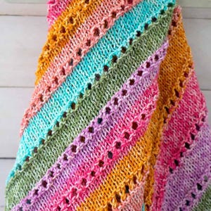 Puede incluir: Una manta de punto de rayas de colores con un patrón de encaje. La manta está hecha con una variedad de colores, incluyendo rosa, naranja, amarillo, verde y azul.