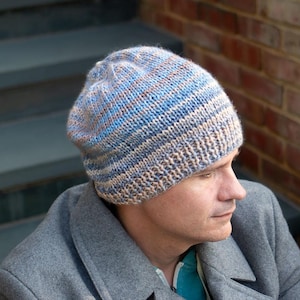 May include: A knitted beanie hat with horizontal stripes in shades of blue, tan, and brown. The hat is worn by a person wearing a gray coat. The hat appears to be handmade.