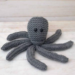May include: A gray knitted octopus with black button eyes. The octopus has eight tentacles and is made of a soft, textured yarn.