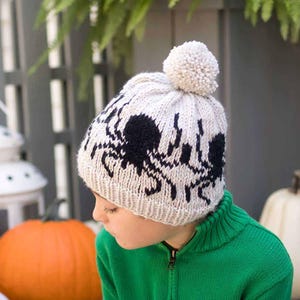 May include: A cream-colored knit beanie featuring a repeating black spider pattern and a matching cream-colored pom-pom on top. The hat has a ribbed brim and is constructed from thick yarn, displaying a textured knit stitch throughout the design.