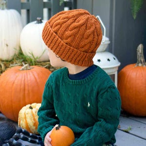 Könnte beinhalten: Ein junges Kind trägt eine gestrickte orangefarbene Mütze mit einem Zopfmuster. Die Mütze sitzt auf seinem Kopf. Das Kind trägt einen grünen Pullover und hält einen kleinen orangefarbenen Kürbis in der Hand. Das Kind sitzt vor einem Haufen Kürbisse.