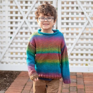 May include: A young person wearing a colorful striped sweater with a crew neck. The sweater has shades of blue, green, pink, and orange. The person is wearing brown pants and glasses.