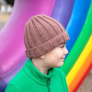 Puede incluir: Un gorro de punto marrón con textura de canalé. El gorro lo lleva puesto una persona de pie frente a un fondo de arcoíris de colores.