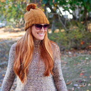 May include: A woman wearing a brown knit beanie with a pom pom on top and brown sunglasses. She is wearing a speckled brown sweater.