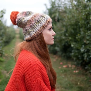 May include: A woman wearing a colorful knitted beanie with a pom-pom on top. The beanie has a mix of orange, brown, and green colors. She is wearing a red sweater and has long red hair.