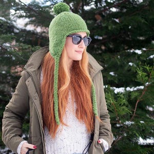 May include: A green knitted hat with a pom pom on top and long ear flaps. The hat is worn by a person with long red hair and sunglasses.