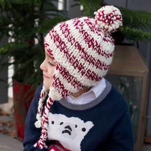 May include: A white and red striped knit hat with ear flaps and a pom pom on top. The hat is worn by a child wearing a blue sweater with a white polar bear design.