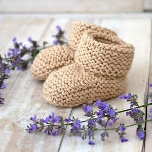 May include: A pair of beige knitted baby booties with a fold-over cuff. The booties are sitting on a white wooden surface with purple flowers.