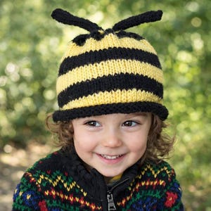 May include: A child wearing a knitted bee hat with yellow and black stripes and antennae. The hat has a black brim. The child is also wearing a colorful sweater with a zipper.
