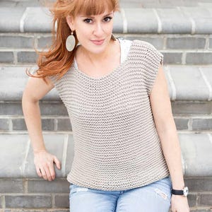 May include: A beige, sleeveless knitted top with a V-neck. The top has a textured, ribbed appearance. The person is wearing blue jeans and a black watch. The background is a brick wall and steps.