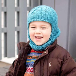 May include: A young child wearing a blue knitted hat with a fold-down flap covering the ears and neck. The hat is worn over a colorful patterned sweater and a brown jacket.