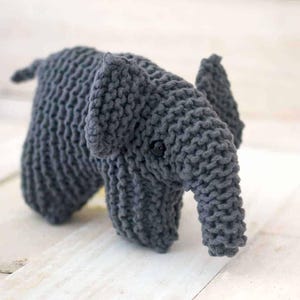 Op de afbeelding: Een handgemaakt grijs gebreid olifantenspeelgoed met een knoopoog en een slurf. De olifant heeft grote oren en een kleine staart. Het speelgoed is gemaakt van zacht, getextureerd garen en staat op een wit oppervlak.