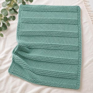 Puede incluir: Una manta tejida rectangular de color verde salvia sólido. La manta presenta un patrón texturizado que consiste en secciones alternas de punto jersey liso y bandas horizontales de filas de punto bobo en relieve.