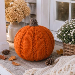 May include: A round, orange, hand-knitted pumpkin decoration featuring a textured, ribbed surface and a small, brown, crocheted stem at the top. The pumpkin is crafted from thick yarn and displays a consistent, dense knit pattern throughout its exterior.