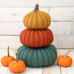 May include: A stack of three knitted pumpkins in autumnal colours: mustard yellow, burnt orange, and forest green. The pumpkins are stacked on a white wooden surface. There are three small orange pumpkins in the foreground.