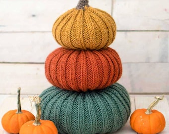 Pode incluir: Uma pilha de três abóboras de tricô em cores outonais: amarelo mostarda, laranja queimado e verde floresta. As abóboras estão empilhadas sobre uma superfície de madeira branca. Há três pequenas abóboras laranja em primeiro plano.