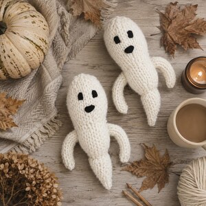 May include: Two off-white knitted ghost figures with black yarn eyes and mouths. Each ghost has a rounded head, two arms extending outward, and a tapered tail-like base. The soft, textured yarn construction creates a simple, rounded shape.