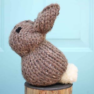 May include: A handmade brown knitted bunny figurine with a fluffy white tail. The bunny has black bead eyes and is sitting on a wooden base. The ears are upright, and the overall design is rustic and charming.