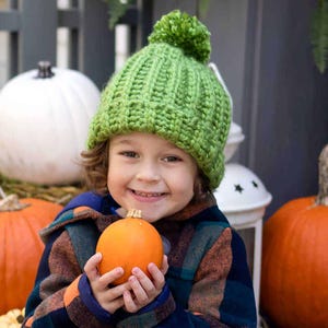 May include: A young child wearing a green knitted hat with a pom pom smiles while holding a small orange pumpkin. The child is standing in front of a background of pumpkins.