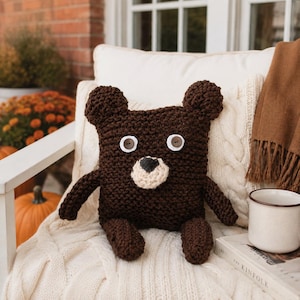 May include: A dark brown square crochet bear pillow featuring two round ears, two white and brown button eyes, and a light beige stitched nose. The bear has two small arms and two small legs attached to the main body, all crafted from a textured yarn.