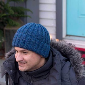 May include: A blue knitted beanie with a ribbed texture. The beanie is worn by a person with a dark jacket.