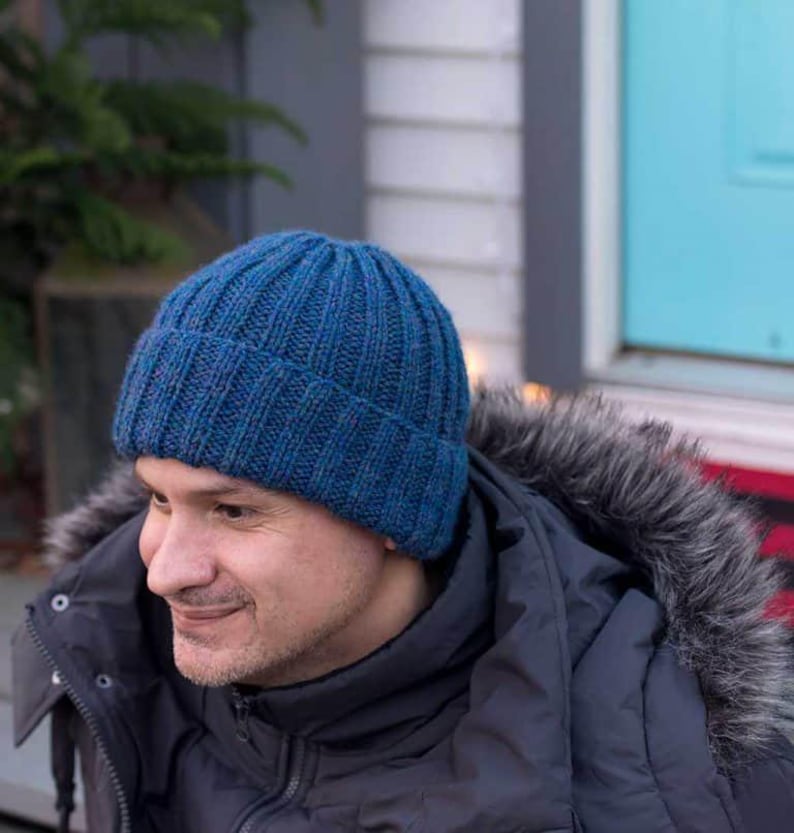 May include: A blue knitted beanie with a ribbed texture. The beanie is worn by a person with a dark jacket.