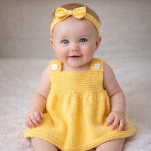 May include: A smiling baby wearing a yellow knitted dress with white buttons on the straps. The baby also wears a matching yellow headband with white polka dots. The dress has a simple, classic design.