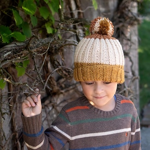 May include: A ribbed knit beanie with a pom-pom in shades of cream, brown, and mustard yellow. The hat is worn by a person wearing a brown sweater with horizontal stripes in orange, blue, and white.