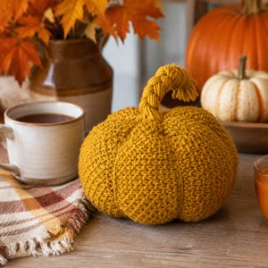May include: A mustard yellow crocheted pumpkin with a thick, braided stem. The pumpkin features a textured, ribbed surface created by the yarn stitches. It is a round, soft-sculpture object designed with a distinct stem that curves slightly at the top.