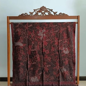 Könnte beinhalten: Ein dunkelbrauner und roter Batikstoff mit einem floralen Muster wird auf einem Holzständer ausgestellt. Der Stoff hat ein traditionelles indonesisches Design.