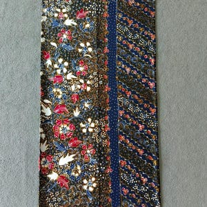 Op de afbeelding: Een rechthoekige batikstof met een bloemenpatroon in rood, wit en blauw op een donkere achtergrond. Het heeft een blauwe rand met witte stippen en een contrasterend patroon. De stof is waarschijnlijk katoen of een vergelijkbaar materiaal.