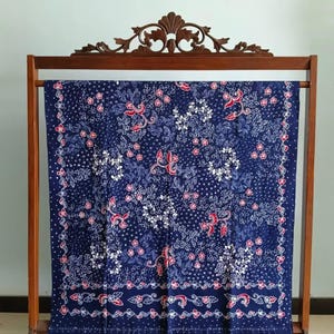 Könnte beinhalten: Ein blauer Batikstoff mit einem weißen und roten Blumenmuster wird auf einem Holzgestell präsentiert. Der Stoff hat einen Rand mit einem sich wiederholenden Blumendesign.