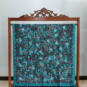 May include: A dark textile with a teal floral pattern and border is displayed on a wooden rack. The fabric features a detailed design of flowers and leaves against a black background, creating a striking contrast. The rack has a decorative top.