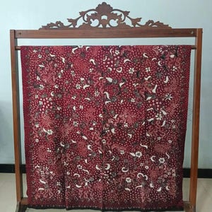 May include: A red batik fabric with a white and black floral pattern displayed on a wooden stand.