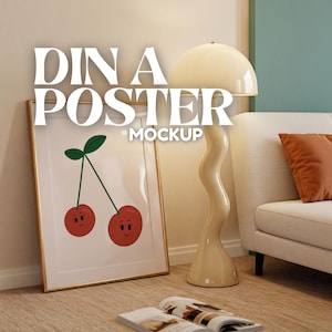 May include: A gold framed poster mockup with a white background and a cartoon illustration of two red cherries with smiling faces. The poster is leaning against a wall next to a beige lamp with a white shade and a white couch with an orange throw pillow.