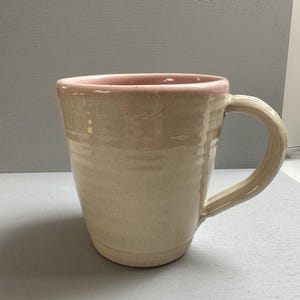 May include: A handmade ceramic mug with a beige exterior and a pink interior rim. The mug has a rounded body, a sturdy handle, and a speckled texture. The mug is designed for hot beverages.