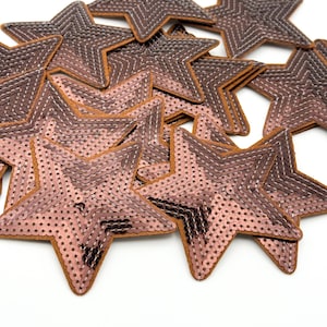 Può includere: Una collezione di toppe a forma di stella. Le toppe sono principalmente di colore oro rosa metallizzato, con un bordo marrone e cuciture bianche. Le stelle sono sparse su uno sfondo bianco, creando un effetto strutturato e decorativo.