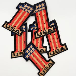 May include: Five red, white, and blue embroidered patches shaped like the number one. Each patch has the word "USA" written on it.